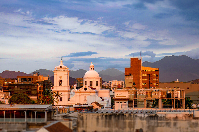 Luftaufnahme vom historischen Herz von Santa Marta bei Dämmerung, überragt von der schneeweißen Kathedrale, deren Turm und Kuppel im weichen Abendlicht warm leuchten. Santa Marta Kolumbien Sonnenuntergang