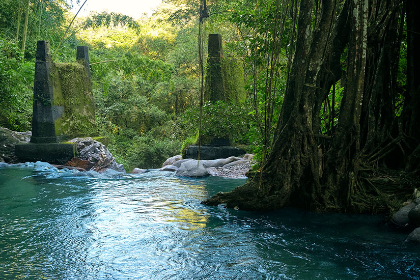 Ein klares, smaragdgrünes Naturbad im Regenwald, das im Vordergrund sanfte Wellen schlägt. Zwei moosbedeckte, quadratische Betonpfeiler ragen wie antike Ruinen aus dem Wasser und der ufernahen Vegetation empor. Santa Marta Kolumbien Povo Azul Wasserfall