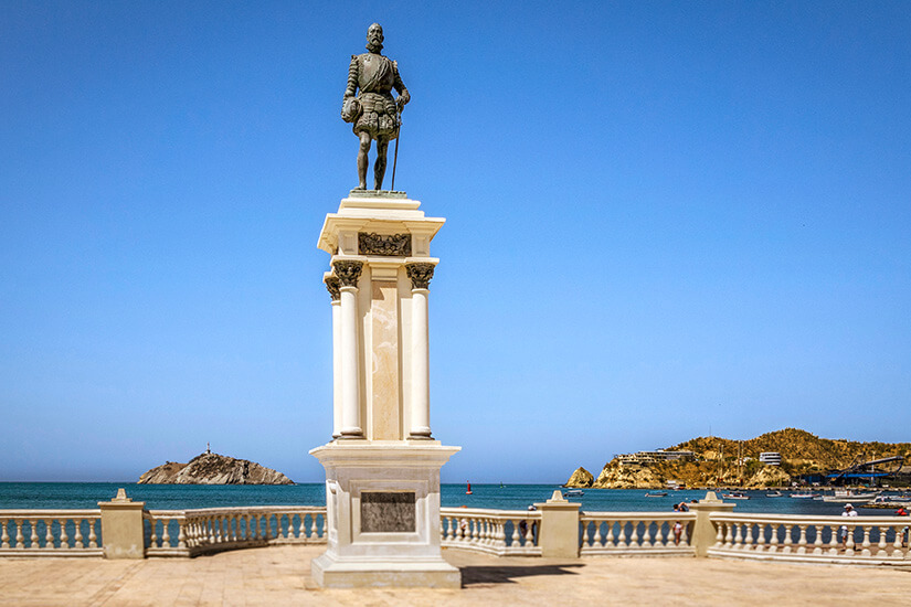 Eine bronzene Statue von Rodrigo de Bastidas, die auf einem hohen, hellbeigen Marmorsockel mit korinthischen Säulenelementen steht. Die Figur blickt in historischer Rüstung mit einem Schwert an der Seite auf das Karibische Meer hinaus. Santa Marta Kolumbien Rodrigo de Bastidas Statue