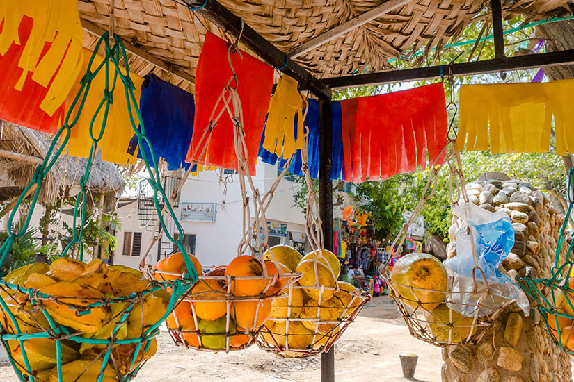 Ein rustikaler Marktstand unter einem Dach aus getrockneten Palmblättern. An einer schwarzen Metallstange hängen bunte Papiergirlanden in Gelb, Blau und Rot, den Nationalfarben Kolumbiens. Darunter befinden sich handgeknüpfte, weiße und grüne Netzkörbe, die mit verschiedenen Früchten gefüllt sind. Santa Marta Kolumbien Obststand