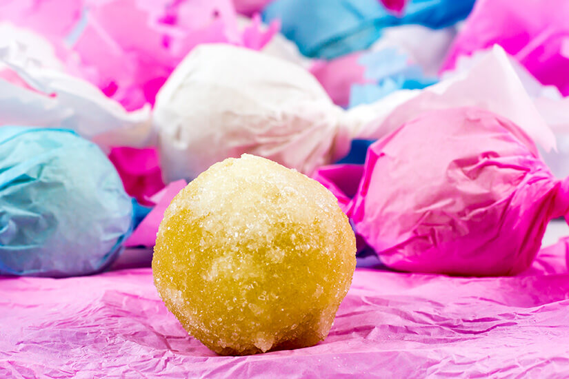 Eine runde Süßigkeit mit zuckerbestreuter Oberfläche liegt im Vordergrund auf pinkem Papier. Im Hintergrund sind weitere Bonbons oder Gebäckstücke in buntem Papier eingewickelt. Souvenirs Sardinien Sospiri