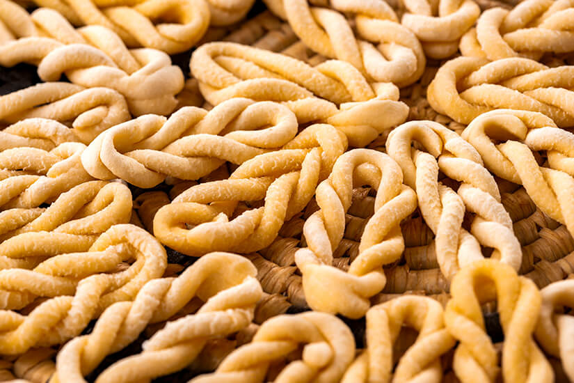 Mehrere handgeformte, gedrehte Pastastücke liegen dicht nebeneinander auf einer geflochtenen Unterlage. Die hellgelbe Farbe und die leicht raue Oberfläche deuten auf frische, ungekochte Nudeln hin. Die gleichmäßigen Schleifenformen wiederholen sich über das gesamte Bild. Sardinien Pasta