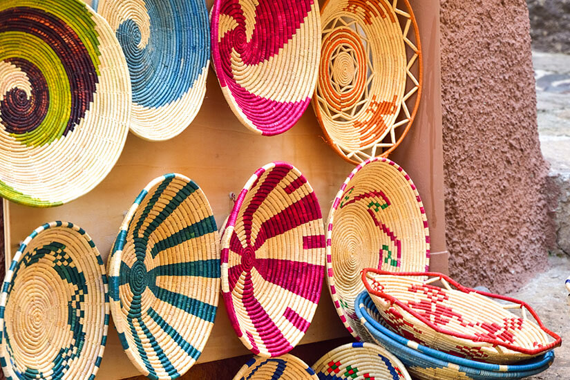 Mehrere runde, handgeflochtene Körbe mit bunten Mustern lehnen an einer Wand. Die Designs bestehen aus spiralförmigen und geometrischen Formen in kräftigen Farben wie Rot, Grün und Blau. Einige Körbe sind übereinandergestapelt. Sardinien Flechtkorb