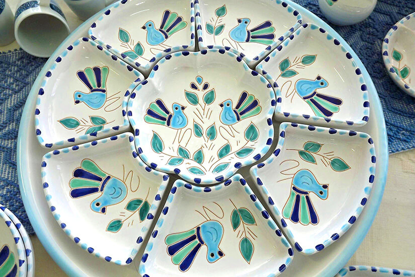 Ein rundes Keramik-Serviertablett mit mehreren unterteilten Fächern ist kunstvoll bemalt. Blaue Vögel und grüne Blätter zieren die einzelnen Segmente sowie die Mitte des Tellers. Souvenirs Sardinien Keramik