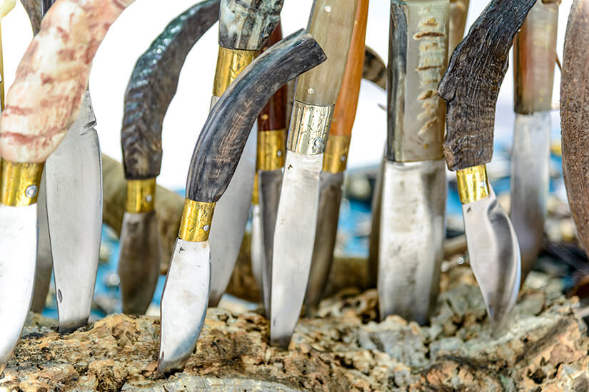 Mehrere Messer mit gebogenen Klingen stecken nebeneinander in einem Stück Holz. Die Griffe bestehen aus unterschiedlichen Materialien wie Holz oder Horn und sind teilweise mit Metallverzierungen versehen. Sardinien Messer