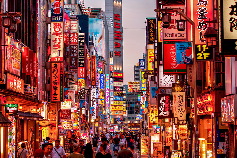 Eine belebte Gasse in Tokio bei Nacht, gesäumt von unzähligen leuchtenden Schildern und Neonreklamen. Das bunte Treiben der Passanten zwischen den eng stehenden Gebäuden fängt die einzigartige Energie dieser Megametropole ein. Städte Japan Tokio