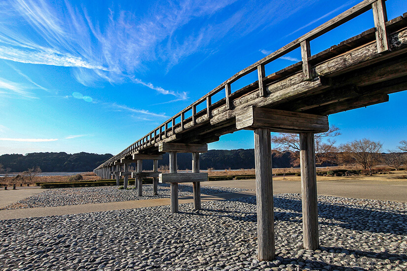 Die Horai-Brücke in Shimada, die längste Holzfußgängerbrücke der Welt, erstreckt sich über ein weites Flussbett unter strahlend blauem Himmel. Die schlichte Holzkonstruktion vor der dramatischen Wolkenkulisse wirkt beinahe meditativ. Städte Japan Shizuoka