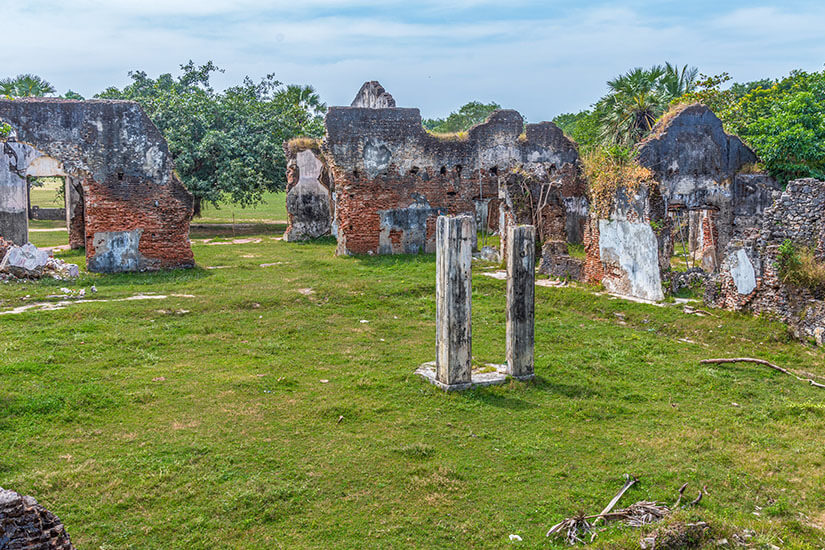 Verfallene Backsteinmauern und verwitterte Säulenstümpfe einer ehemaligen Kolonialanlage erheben sich gespenstisch auf einer gepflegten Rasenfläche, während tropische Vegetation im Hintergrund die Ruinen umrahmt. Jaffna Sri Lanka Fort