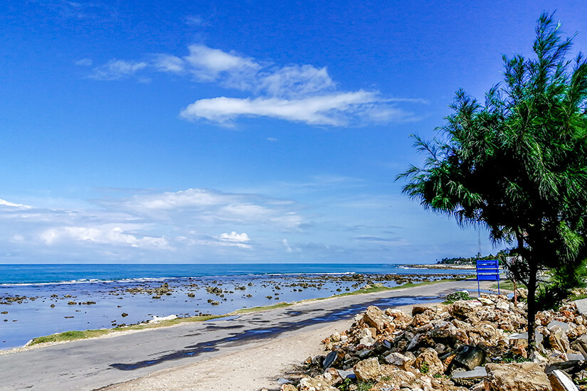 Eine asphaltierte Küstenstraße verläuft entlang eines felsigen Strandes bei Ebbe, wo Korallenreste und Steine aus dem seichten Wasser ragen. Jaffna Sri Lanka Point Pedro