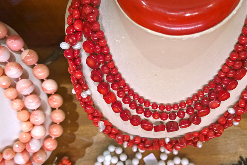 Mehrere Halsketten aus roten und rosafarbenen Perlen sind auf einer Schmuckbüste arrangiert. Die Perlen variieren in Größe und Form. Daneben liegen weitere Schmuckstücke aus ähnlichen Materialien. Souvenirs Sardinien Schmuck
