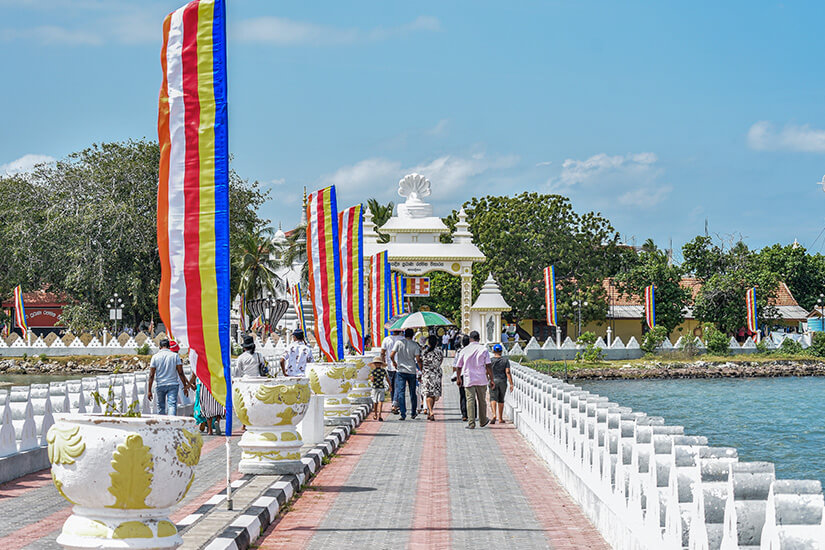 Eine von bunten buddhistischen Flaggen gesäumte Brücke führt über Wasser zu einem weißen Tempeltor, wahrscheinlich in Sri Lanka. Mehrere Besucher schlendern in der Mittagssonne über den gepflasterten Weg in Richtung des Heiligtums. Nagadeepa Purana Vihara