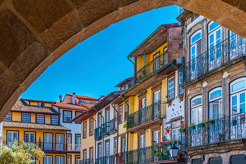 Blick durch einen Steinbogen auf farbenfrohe historische Häuserzeilen in der Altstadt von Guimarães unter strahlend blauem Himmel. Die mehrstöckigen Gebäude haben gelbe und weiße Fassaden sowie traditionelle Balkone mit schwarzen Schmiedeeisengittern und bunten Blumenkästen. Guimaraes Portugal Altstadt