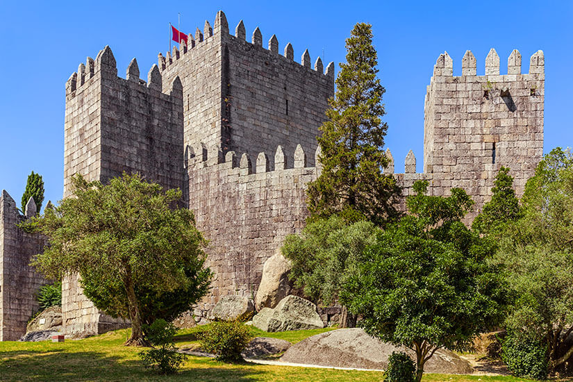 Die mittelalterliche Burg von Guimarães mit ihren markanten, quadratischen Zinnentürmen aus grauem Granitstein. Die Festung steht auf einem grünen Hügel mit Felsen und Bäumen unter klarem blauen Himmel; auf dem höchsten Turm weht eine rote Flagge. Guimaraes Portugal Castelo