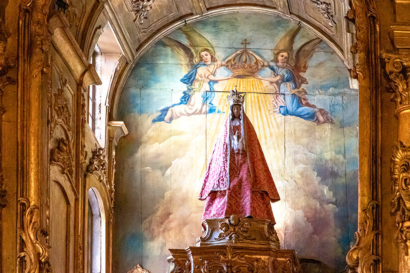 Detailansicht des vergoldeten Hauptaltars in der Igreja da Nossa Senhora da Oliveira in Guimarães. Eine Statue der Gottesmutter Maria steht vor einem hellen Wandgemälde, das zwei schwebende Engel zeigt, die eine Krone über ihr Haupt halten. Die Szenerie ist von prunkvollen, barocken Goldverzierungen und geschnitzten Säulen eingerahmt. Guimaraes Portugal Nossa Senhora da Oliveira