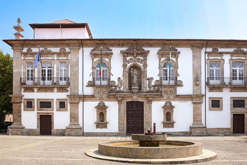 Vorderansicht des barocken Rathauses von Guimarães, dem ehemaligen Bischofspalast, an einem sonnigen Tag. Die weiße Fassade ist reich mit Steinschnitzereien, kunstvollen Fensterumrahmungen, Balkonen mit schmiedeeisernen Geländern und einer zentralen Figurennische verziert. Vor dem Gebäude, auf dem gepflasterten Platz, befindet sich ein runder Steinbrunnen. Links und rechts wehen Flaggen. Guimaraes Portugal Convento de Santa Clara
