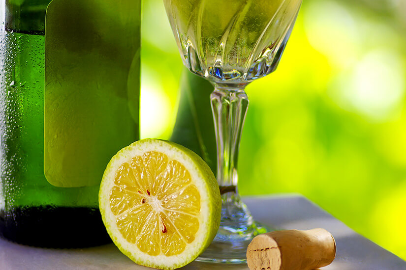 Nahaufnahme einer beschlagenen grünen Weinflasche, einer aufgeschnittenen Limettenhälfte und einem Kristallglas mit Vinho Verde auf einem Marmortisch. Ein Korken liegt daneben; der Hintergrund ist ein unscharfes, leuchtend grünes Blätterdach. Guimaraes Portugal Vinho Verde