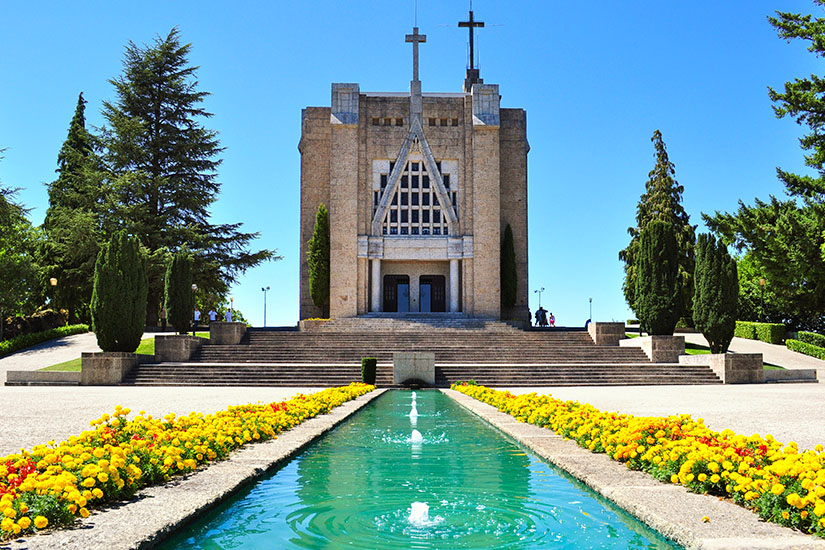 Moderne Steinkirche auf dem Berg Penha mit einem langen Wasserbecken und gelben Blumenrabatten im Vordergrund unter blauem Himmel. Guimaraes Portugal Penha Kapelle