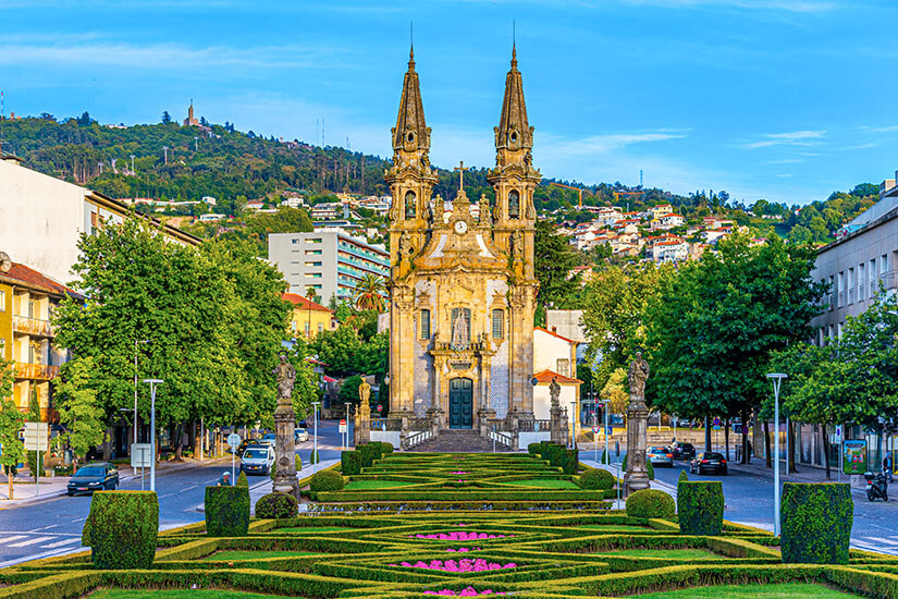 Blick über eine kunstvoll gestaltete Gartenanlage mit geometrischen Hecken auf die barocke Kirche mit ihren zwei markanten Türmen am Ende des Platzes. Guimaraes Portugal Kirche