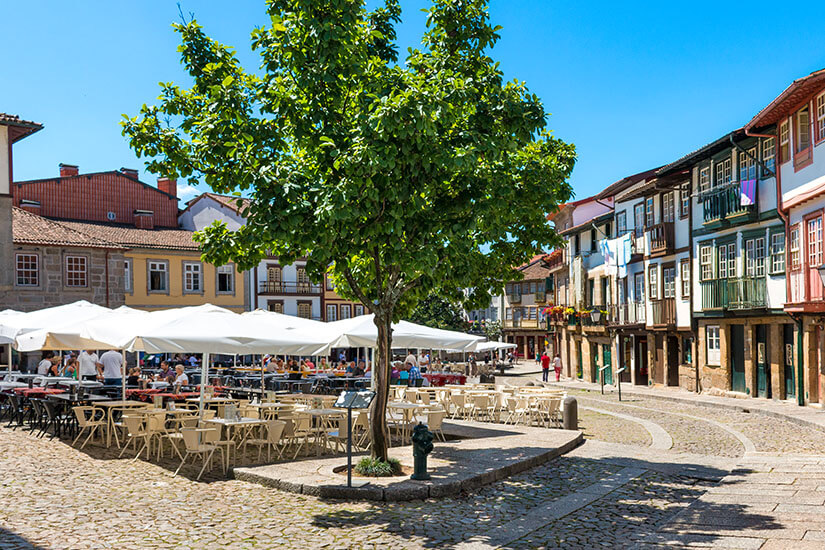 Belebter Platz mit zahlreichen weißen Sonnenschirmen eines Restaurants unter einem großen grünen Baum, umgeben von historischen Häuserzeilen. Guimaraes Portugal Restaurant
