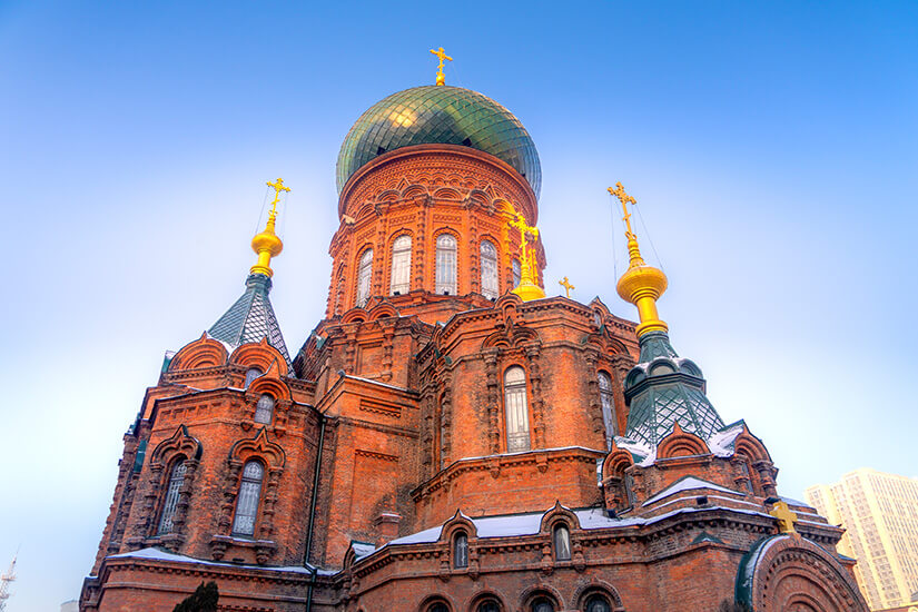 Eine große Kirche aus rotem Backstein mit einer grünen Kuppel und mehreren goldenen Kreuzen erhebt sich gegen den Himmel. Die Architektur wirkt reich verziert mit Rundbögen, Türmchen und ornamentalen Details. Teile des Gebäudes sind leicht mit Schnee bedeckt. Harbin Kathedrale