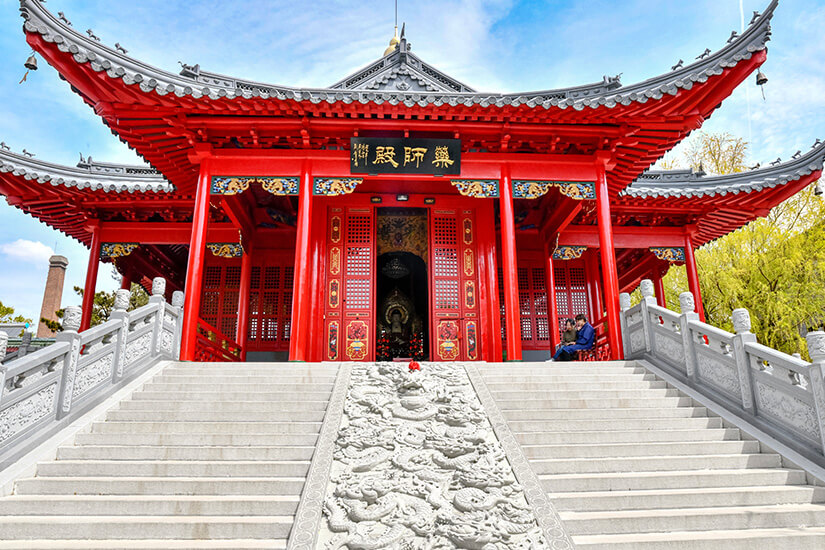Ein auffällig roter Tempel mit geschwungenem Dach und reich verzierten Details steht am oberen Ende einer breiten Treppe. Die Stufen führen zu einem Eingang mit kunstvollen Türen und dekorativen Ornamenten. An den Seiten der Treppe befinden sich steinerne Geländer, und im Inneren des Gebäudes ist eine Statue zu erkennen. Harbin Jile Tempel