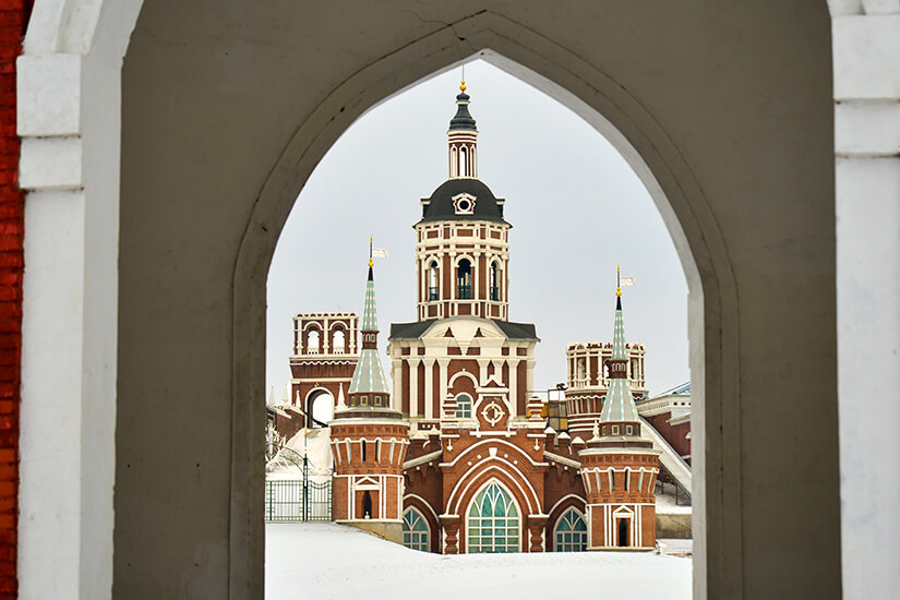 Durch einen großen Torbogen blickt man auf ein prächtiges Gebäude mit roten Ziegeln und hellen Verzierungen. Mehrere Türme mit spitzen Dächern und ein zentraler Turm mit Uhr prägen die Architektur. Der Boden ist mit Schnee bedeckt. China Volga Manor