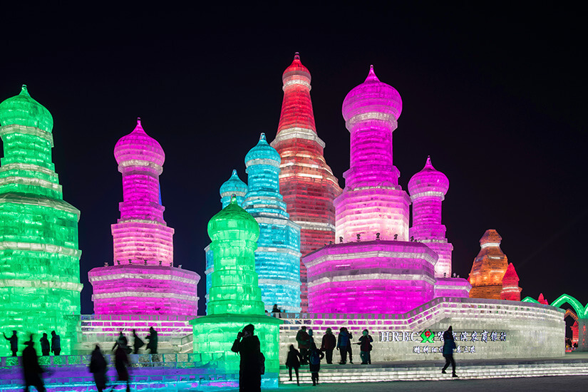 Mehrere große Türme aus Eis sind nachts bunt beleuchtet und erinnern an Pagoden. Die Konstruktionen strahlen in kräftigen Farben wie Grün, Pink und Blau. Im Vordergrund stehen Besucher, die die leuchtenden Eisskulpturen betrachten. Harbin Eisfestival