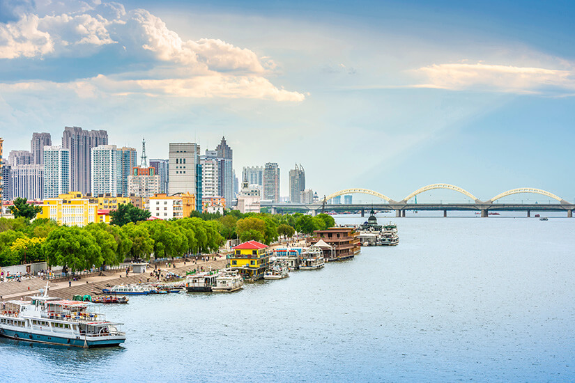 Eine breite Uferpromenade mit Bäumen verläuft entlang eines großen Flusses. Mehrere Ausflugsboote und Anlegestellen liegen am Wasser, während dahinter zahlreiche Hochhäuser eine dichte Skyline bilden. In der Ferne überspannt eine lange Brücke mit mehreren Bögen den Fluss. Harbin Fluss
