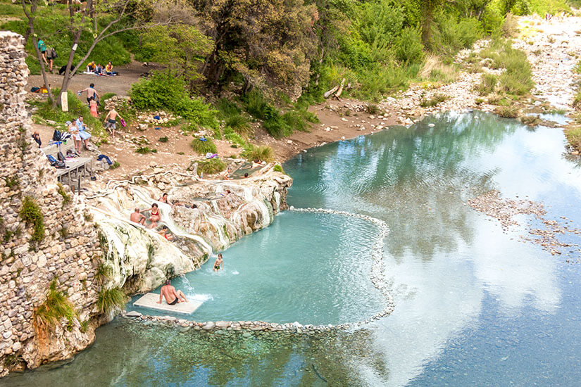 Ein natürliches Thermalbecken mit hellen Kalkablagerungen liegt direkt neben einem Fluss. Mehrere Menschen baden im warmen Wasser oder sitzen am Rand der Felsen. Rundherum befinden sich Bäume, Steine und ein kleiner Uferbereich, auf dem weitere Besucher entspannen. Heiße Quellen Toskana Bagni di Petriolo
