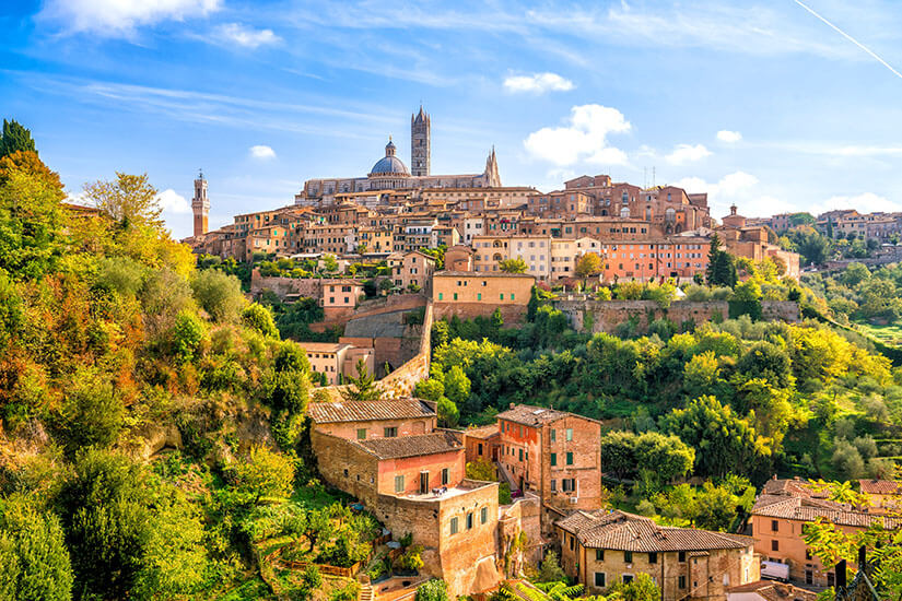Eine historische Stadt liegt auf einem Hügel und besteht aus dicht aneinandergereihten Gebäuden in warmen Erdtönen. Über der Altstadt ragen eine große Kuppel und ein hoher Turm einer Kathedrale heraus. Im Vordergrund befinden sich grüne Hügel, Bäume und einzelne Häuser entlang eines Abhangs. Heiße Quellen Toskana Siena