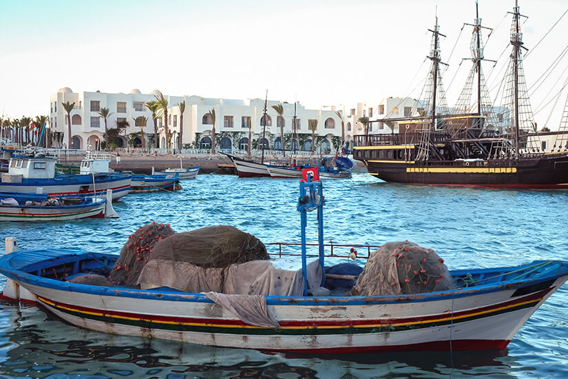 Ein traditionelles tunesisches Fischerboot mit Netzen im Hafen von Houmt Souk vor weißen Gebäuden und einem hölzernen Ausflugsschiff im Hintergrund. Houmt Souk Tunesien Hafen