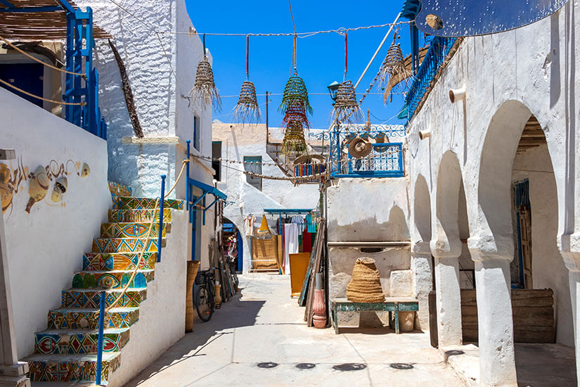 Eine enge, sonnige Gasse in der Altstadt von Houmt Souk mit weiß getünchten Gebäuden, blauen Akzenten, einer kunstvoll bemalten Treppe und hängenden geflochtenen Lampen unter blauem Himmel. Houmt Souk Tunesien Altstadt