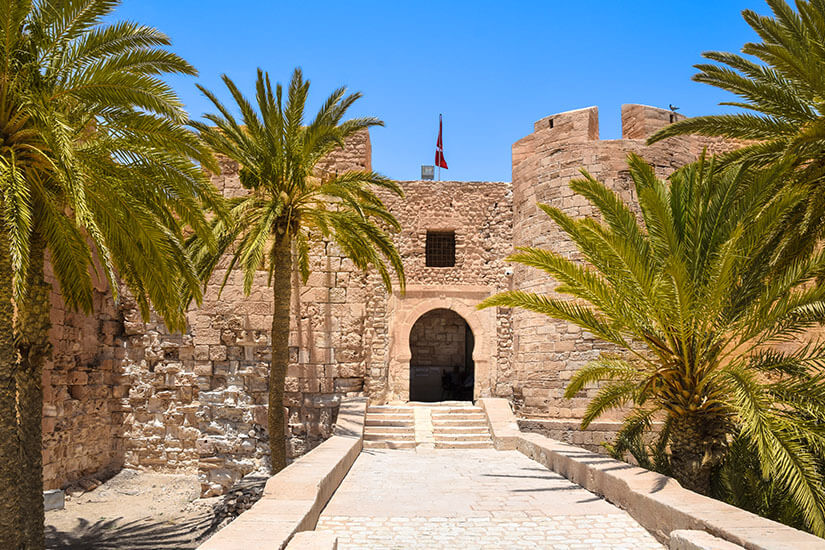 Die historische Festung Borj el Kebir aus hellem Sandstein in Houmt Souk mit einem steinernen Eingangsbereich, flankiert von grünen Palmen unter strahlend blauem Himmel. Houmt Souk Tunesien Borj el Kebir