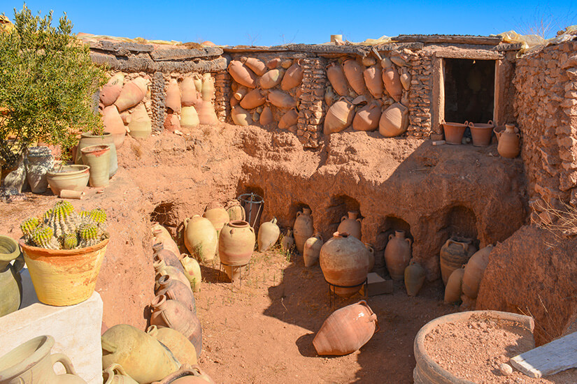 Eine traditionelle Töpferwerkstatt im Freien in Guellala mit zahlreichen Tonkrügen und Amphoren, die in Nischen einer Lehmwand lagern oder im sandigen Boden stehen. Houmt Souk Tunesien Guellala