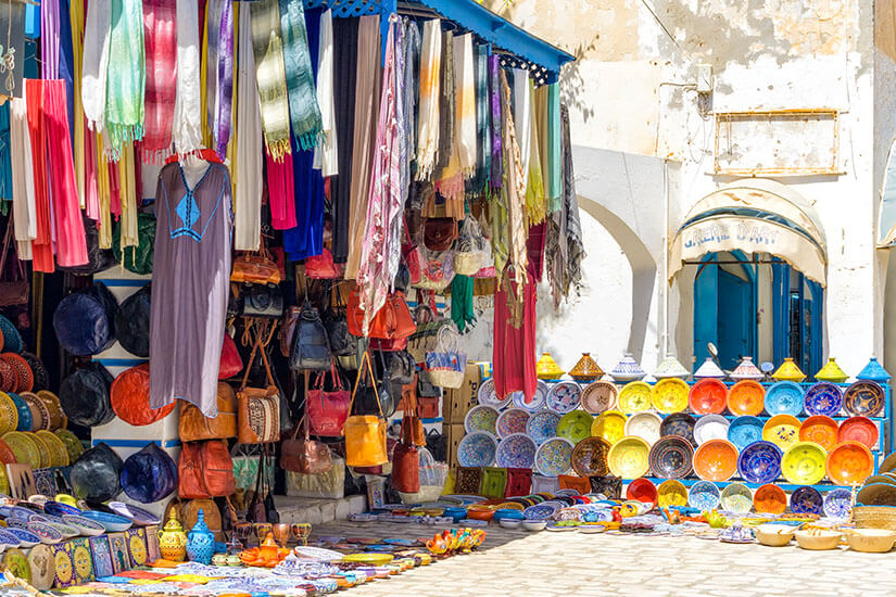 Ein farbenfroher Marktstand in Houmt Souk mit hängenden bunten Stoffen, Ledertaschen und einer großen Auswahl an handbemalter tunesischer Keramik in leuchtenden Gelb und Blau Tönen vor einer weißen Fassade. Houmt Souk Tunesien Markt