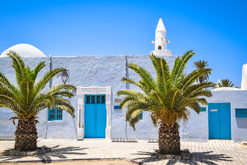 Ein traditionelles, weiß getünchtes Gebäude mit strahlend blauen Holztüren und Fenstergittern auf Djerba, flankiert von zwei großen Palmen unter einem tiefblauen, wolkenlosen Himmel. Rechts ist ein weißes Minarett sichtbar. Houmt Souk Tunesien Moschee