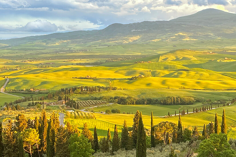 Sanfte, weitläufige Hügel mit grünen und goldgelben Feldern prägen eine typische mediterrane Kulturlandschaft. Zypressenreihen, kleine Straßen und vereinzelte Häuser verteilen sich über die Landschaft. Im Hintergrund erheben sich Berge unter einem Himmel mit dichten Wolken. Italien Val d'Orcia