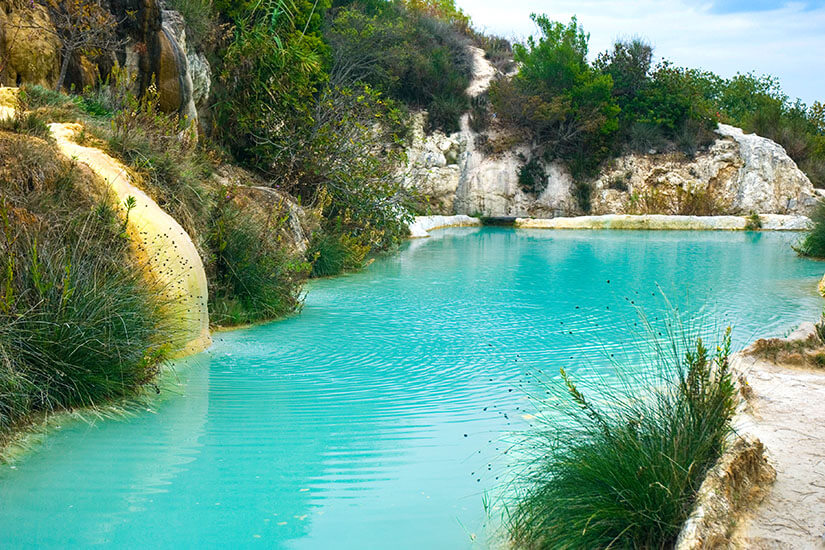 Ein türkisfarbenes Wasserbecken liegt zwischen hellen Kalksteinfelsen und dichter Vegetation. Am Rand wachsen Gräser und Sträucher, während sich ein schmaler Pfad entlang des Wassers zieht. Heiße Quellen Toskana Bagni Vignoni