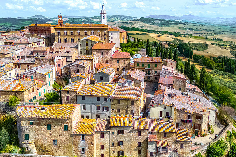 Eine dicht bebaute historische Ortschaft mit vielen Steinhäusern und orangefarbenen Ziegeldächern liegt auf einem Hügel. Über den Gebäuden ragen eine Kirche und ein Turm heraus. Dahinter erstreckt sich eine weite Hügellandschaft mit Feldern, Zypressen und sanften Bergen. Italien Pienza
