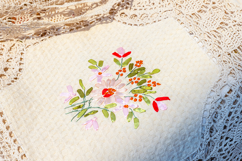 Ein cremefarbenes Textil mit einem fein gestickten Blumenstrauß aus Gänseblümchen, roten Blüten und grünen Blättern liegt auf einer Häkelspitzendecke. Montenegro Souvenirs Spitze