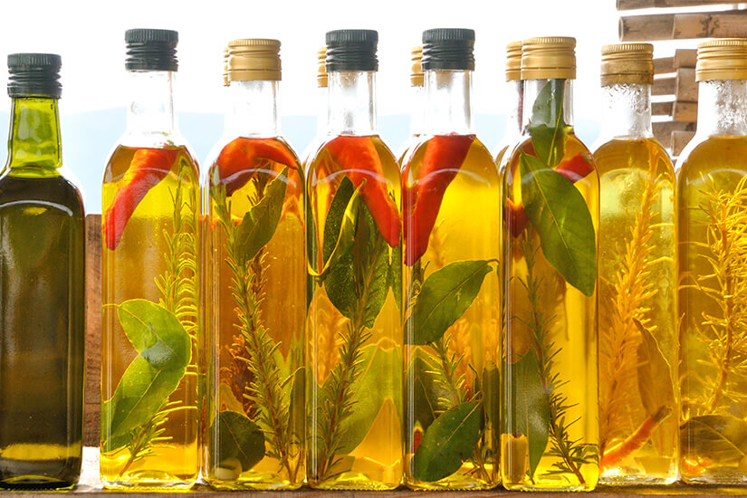 Mehrere Glasflaschen mit aromatisierten Ölen, gefüllt mit Kräutern wie Rosmarin und Lorbeerblättern sowie roten Chilischoten, stehen nebeneinander im Gegenlicht. Montenegro Souvenirs Oel