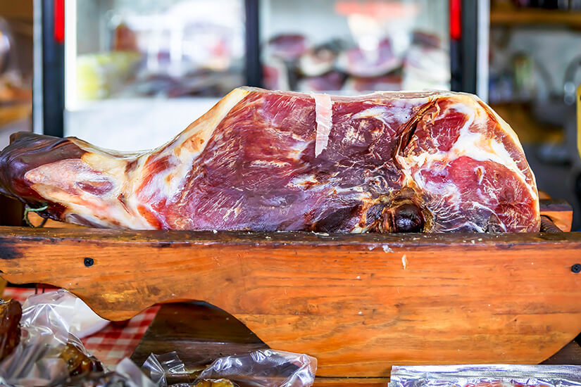 Eine ganze luftgetrocknete Schinkenkeule liegt auf einem hölzernen Bock. Die dunkelrote Fleischfarbe und der charakteristische Fettrand deuten auf ein hochwertiges Produkt hin. Montenegro Souvenirs Schinken
