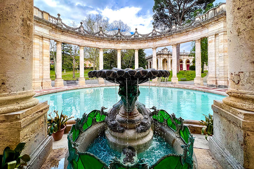 Ein kunstvoller Brunnen sprudelt in einem runden Wasserbecken in der Mitte einer eleganten Anlage. Dahinter verläuft eine halbkreisförmige Kolonnade aus hellen Säulen, die den Platz umrahmt. Quellen Toskana Montecatini Terme