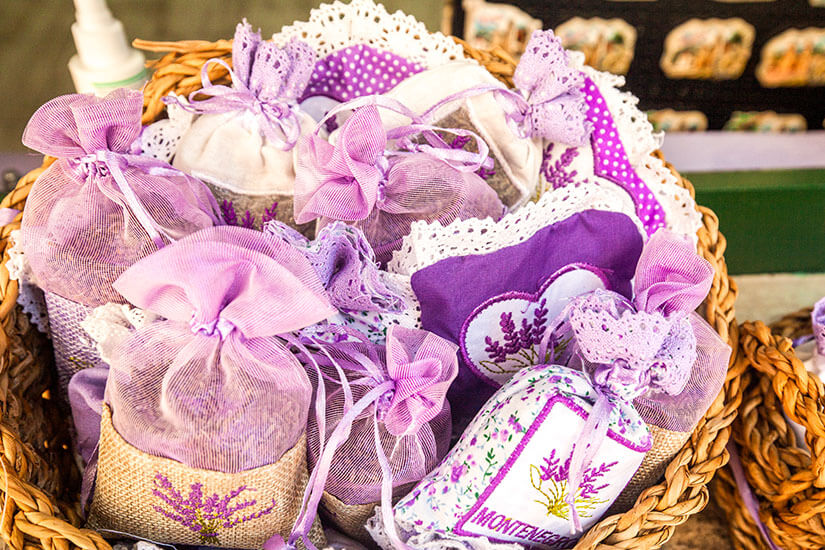 Ein geflochtener Korb ist gefüllt mit hübsch dekorierten Lavendelsäckchen in verschiedenen Lila- und Beigetönen, teilweise mit Spitzenbesatz und Schleifchen verziert. Montenegro Souvenirs Lavendel