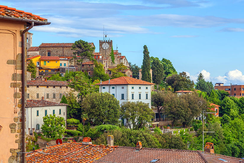 Eine kleine historische Ortschaft mit Steinhäusern und roten Ziegeldächern liegt auf einem grünen Hügel. Zwischen Bäumen ragt ein Turm mit einer großen Uhr über die Gebäude hinaus. Die Umgebung ist von üppiger Vegetation und weiteren Häusern am Hang geprägt. Italien Montecatini Alto