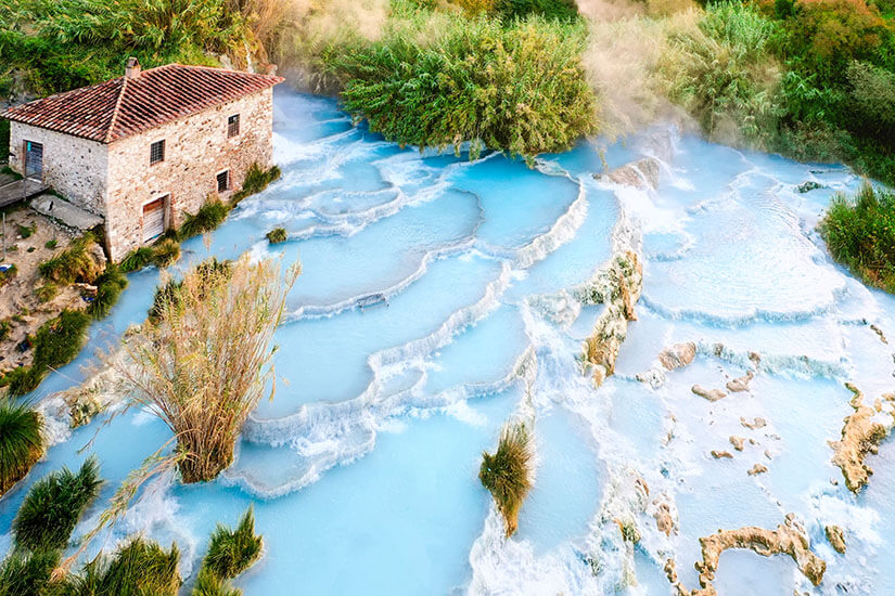 Terrassenförmige Kalkbecken mit hellblauem, dampfendem Wasser erstrecken sich in einer natürlichen Landschaft. Am Rand steht ein kleines Steinhaus mit rotem Ziegeldach. Rundherum wachsen dichte Büsche und Gräser. Termi di Saturnia