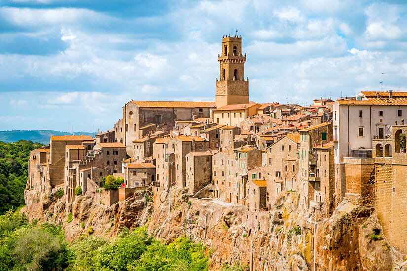 Eine dicht bebaute historische Stadt aus hellen Stein- und Ziegelhäusern steht auf einer steilen Felskante. Über den Gebäuden ragt ein hoher Turm einer Kirche oder eines Rathauses empor. Unterhalb der Häuser fällt der Felsen abrupt zu einer grünen Landschaft ab. Italien Pitigliano