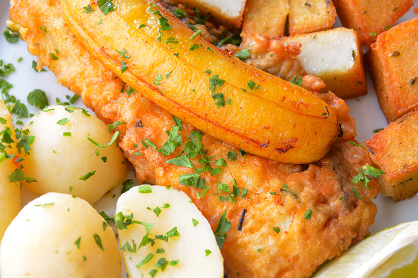 Ein Teller mit einem gebratenen Fischfilet ist mit Kräutern bestreut und mit einer gegrillten Banane belegt. Daneben liegen gekochte Kartoffeln sowie geröstete Maiswürfel. Eine Zitronenscheibe ergänzt das Gericht als Garnitur. Madeira Essen