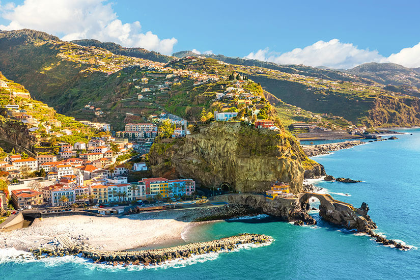 Eine Küstenstadt mit bunten Häusern liegt am Fuß eines steilen Felsens direkt am Meer. Ein kleiner Strand und eine geschwungene Brücke befinden sich am Wasser, während darüber Terrassen und Gebäude den Hang hinaufreichen. Im Hintergrund steigen grüne Berge mit verstreuten Häusern an. Ponta do Sol Stadt