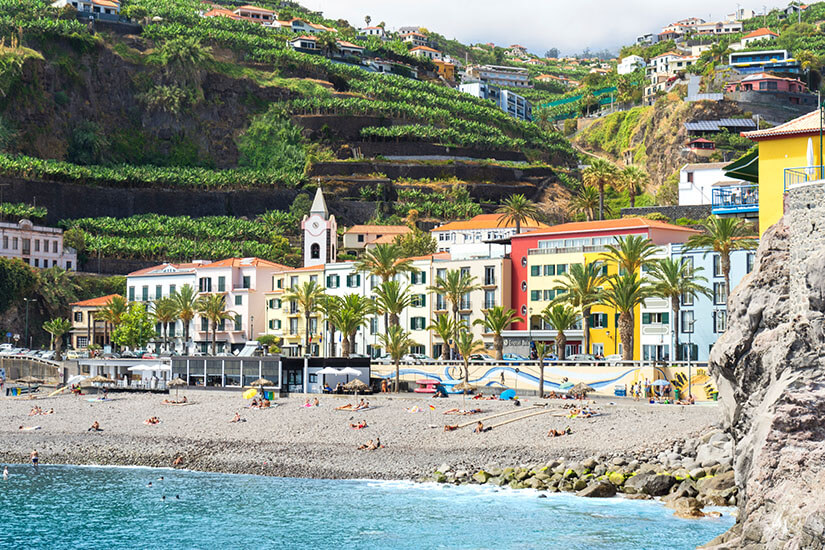 Ein Kiesstrand liegt vor einer Reihe farbenfroher Häuser und Palmen entlang der Uferpromenade. Menschen entspannen am Strand oder liegen in der Sonne nahe dem Wasser. Dahinter steigen steile, grüne Hänge mit Terrassenfeldern und verstreuten Häusern an. Praia da Ponta do Sol